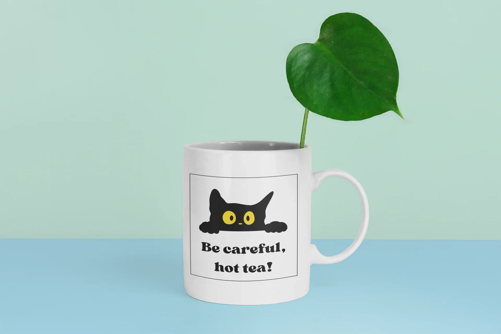 White mug with black cat and yellow eyes, 'Be careful, hot tea!' next to green leaf