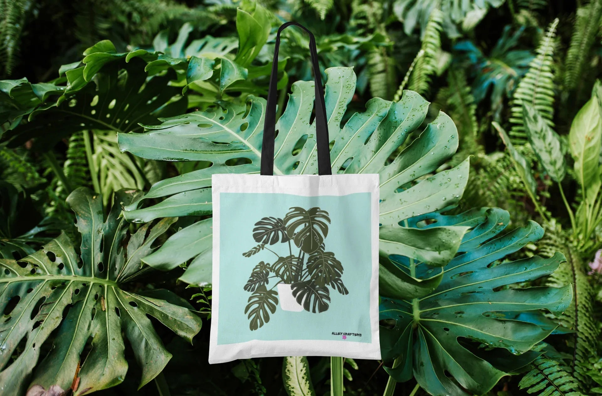White tote bag with black handles, featuring a green monstera leaf design
