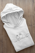 White unisex hoodie folded on wood floor; line-art wedding rings graphic with text on front.