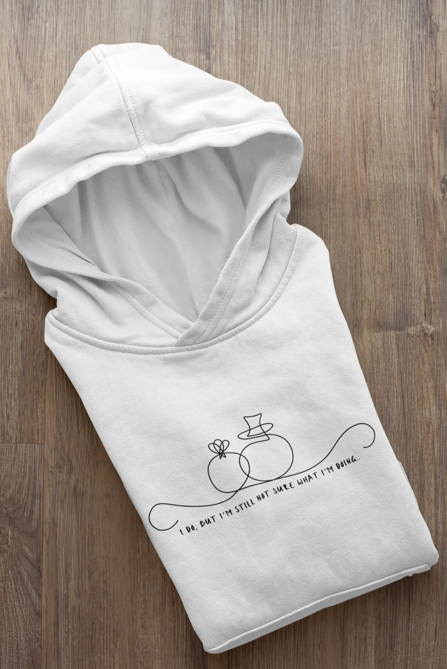 White unisex hoodie folded on wood floor; line-art wedding rings graphic with text on front.