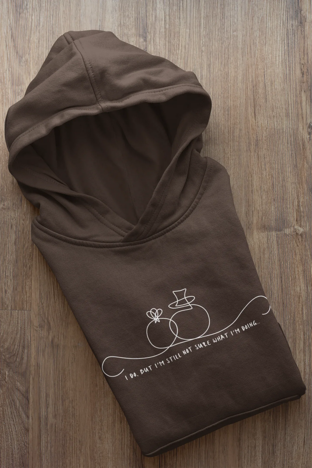 Dark brown unisex hoodie folded flat; line-art wedding rings graphic with text on front.