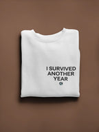 White crewneck sweatshirt with front text graphic “I SURVIVED ANOTHER YEAR” and small icon