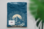 Teal T-shirt with mountain sunrise “RISE ABOVE” logo, folded package view