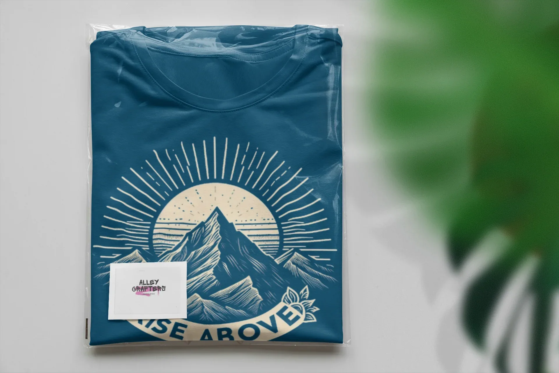 Teal T-shirt with mountain sunrise “RISE ABOVE” logo, folded package view