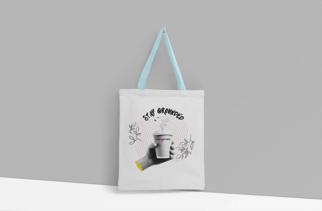 White canvas tote bag with light blue handles and Stay Grounded coffee cup graphic