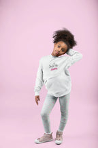 Kids white hoodie with hood and Arabic name print, worn with light patterned leggings.