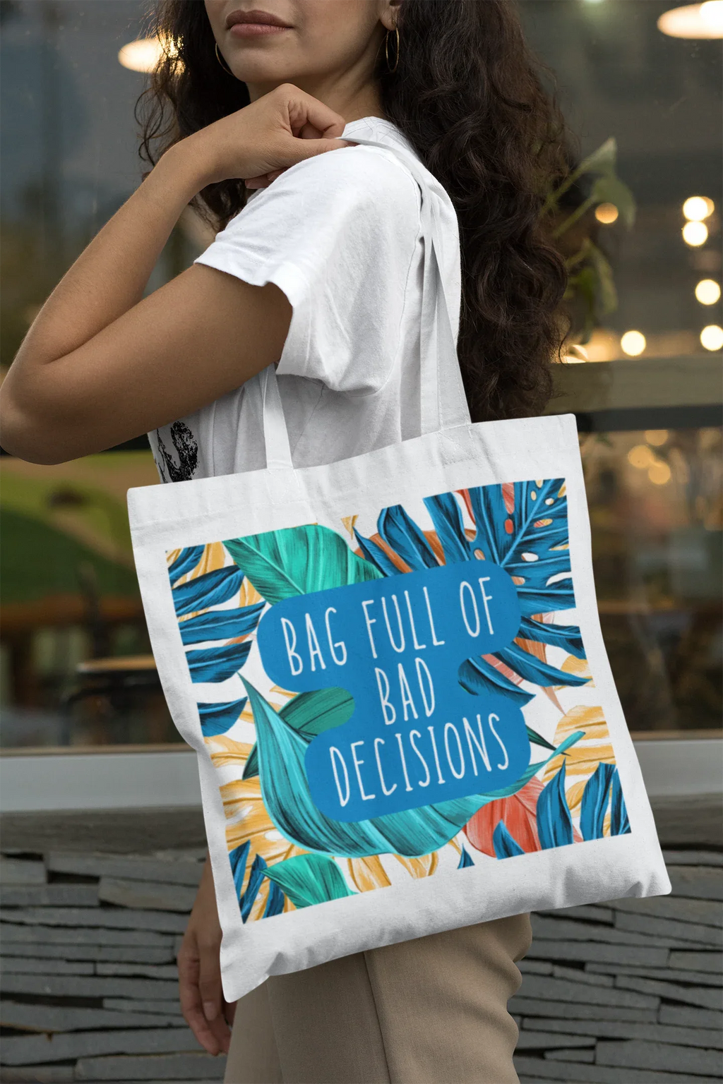 White tote bag with teal circular text design and tropical leaf pattern on front.