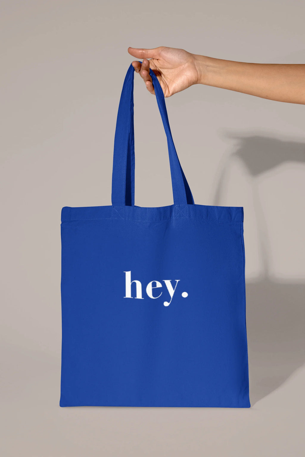 Blue cotton tote bag held by hand with custom text "hey."