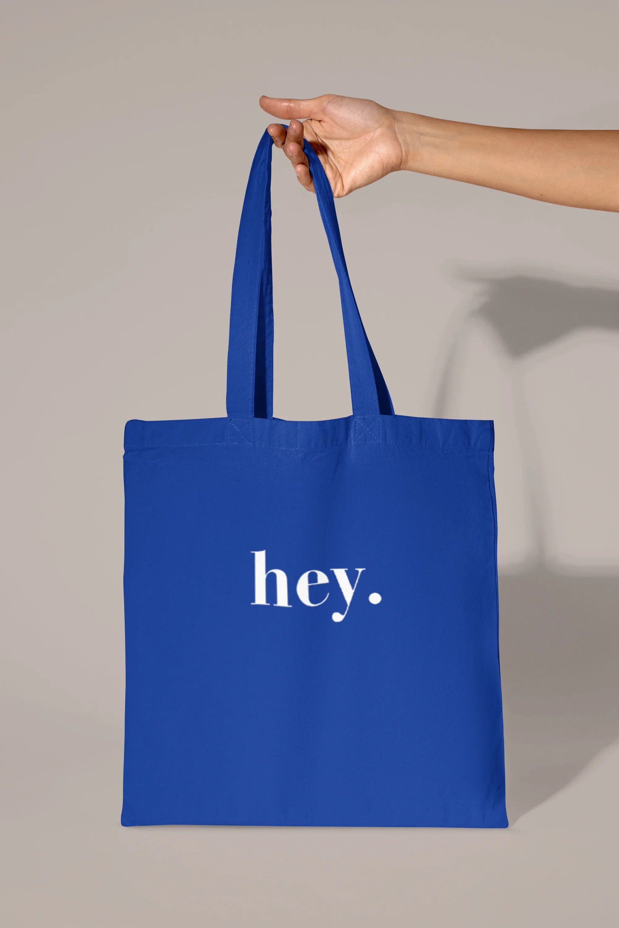 Blue cotton tote bag held by hand with custom text "hey."