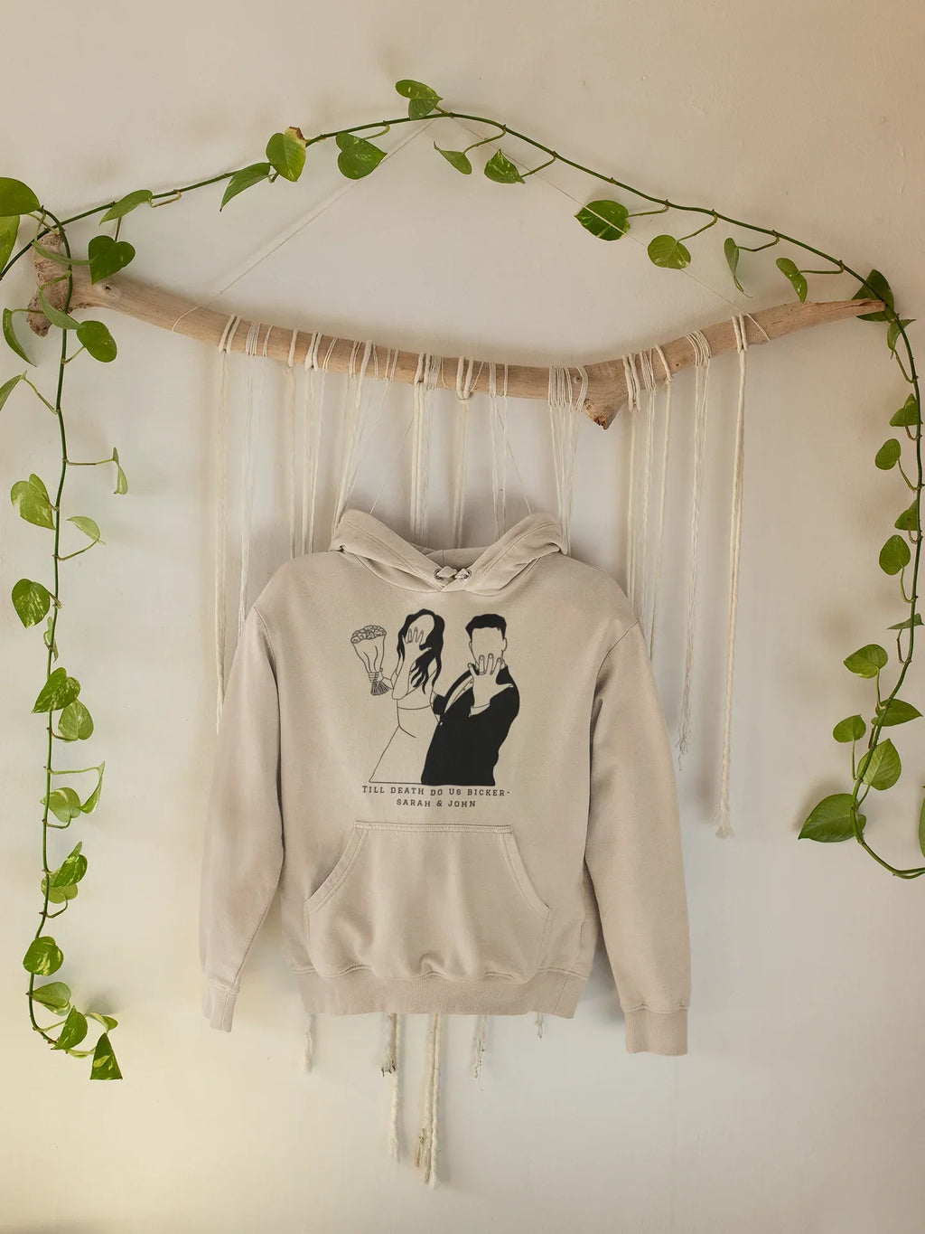 Light-beige hoodie hanging with front couple graphic and text, hood and pouch pocket.