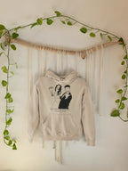 Light-beige hoodie hanging with front couple graphic and text, hood and pouch pocket.