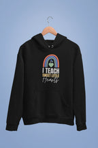 Black unisex hoodie on hanger with front pocket and chest graphic: rainbow heart “I TEACH”