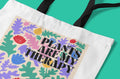 Close-up of a white tote bag panel with “PLANTS ARE MY THERAPY” over leafy floral print