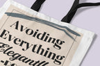 White canvas tote bag close-up with “Avoiding Everything Elegantly” printed in dark