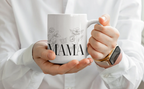 White ceramic coffee mug with 'MAMA' text and floral line art in hands