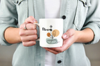 White ceramic mug with Flora name and floral pot graphic, held in hands