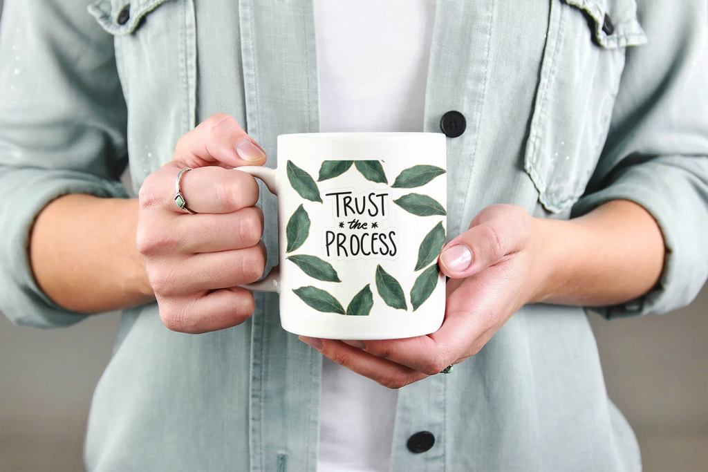 White coffee mug with green leaf wreath and “Trust the Process” held in hands