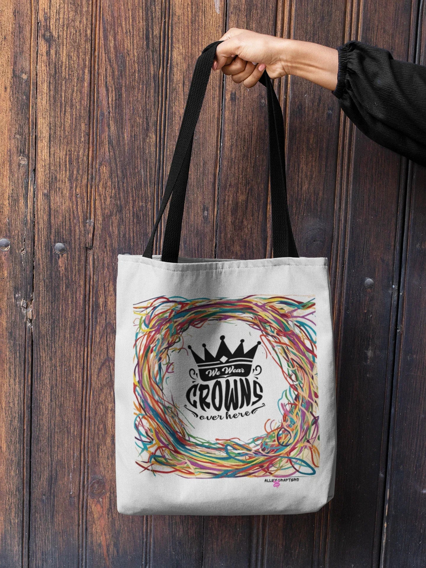 White tote bag held by hand, featuring black crown emblem with multicolor scribble wreath