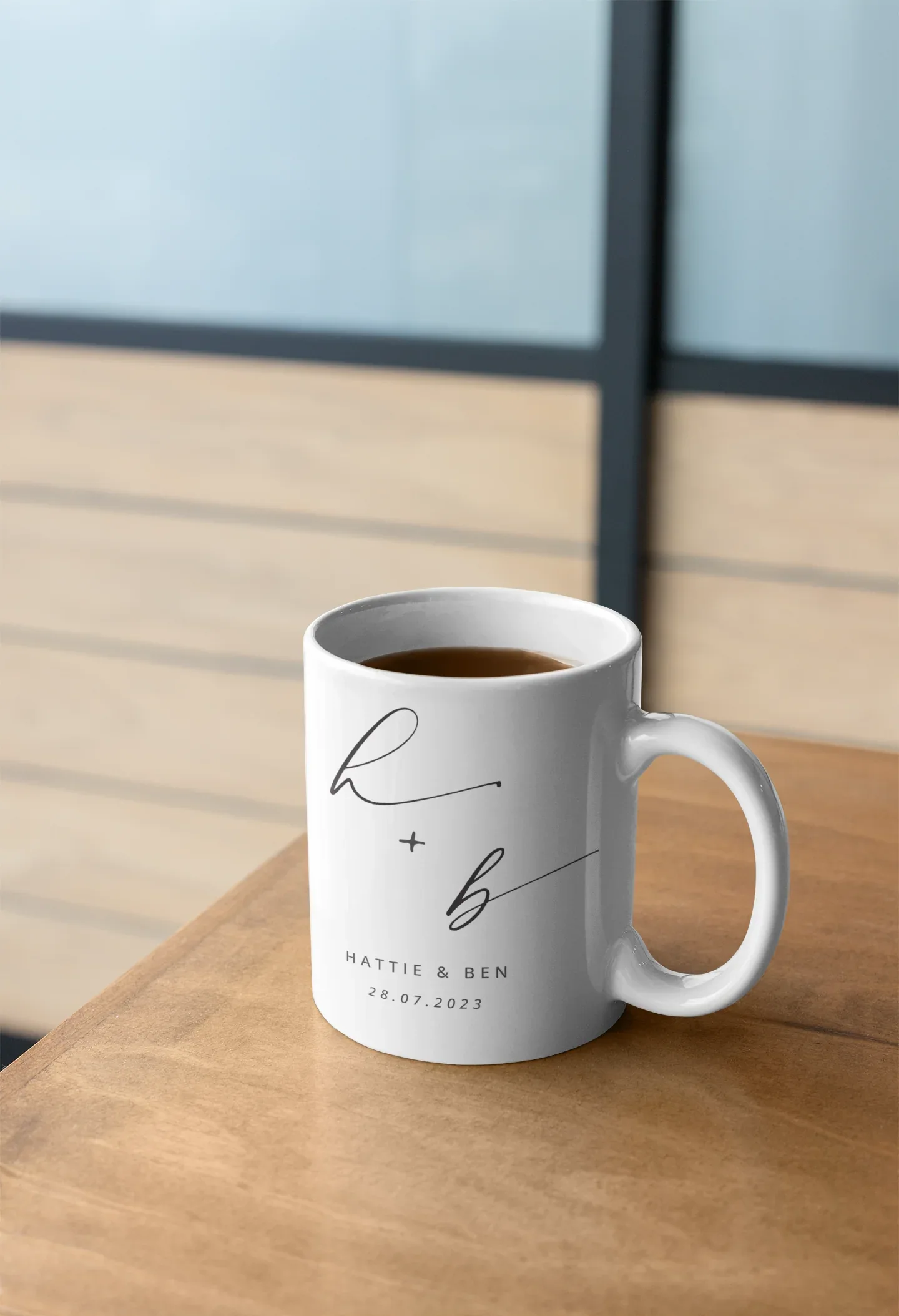 White ceramic mug with monogram h + b and date 28.07.2023, on wooden table.
