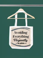 White canvas tote bag hanging, printed "Avoiding Everything Elegantly" text