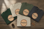Multiple t-shirts stacked in white, green, and charcoal, each with Seasoned wood-slice logo