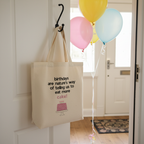 Natural canvas tote bag with birthday quote and pink cake graphic, hanging by a door