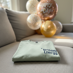 Seafoam T-shirt with Birthday Energy+ graphic, folded on couch