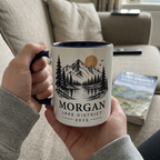 Person holding a dark blue Morgan Lake District 2025 mountain mug on a sofa