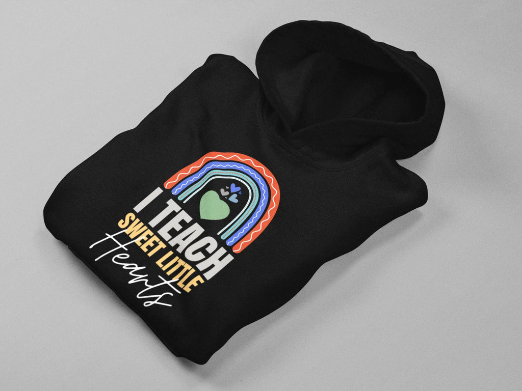 Black unisex hoodie lying flat with front “I TEACH SWEET LITTLE Hearts” rainbow heart print