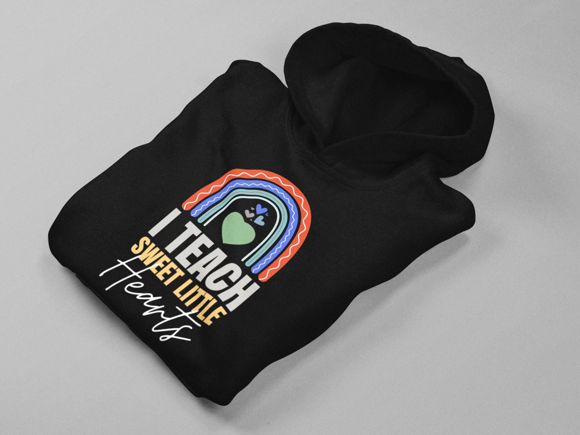 Black unisex hoodie lying flat with front “I TEACH SWEET LITTLE Hearts” rainbow heart print