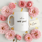 White mug with 'she said yes!' text and heart icon, with pink floral decor.
