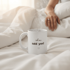 White coffee mug with 'she said yes!' text and small heart icon on bed.