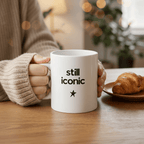 Ceramic coffee mug held in hand, printed “still iconic” with a star on white