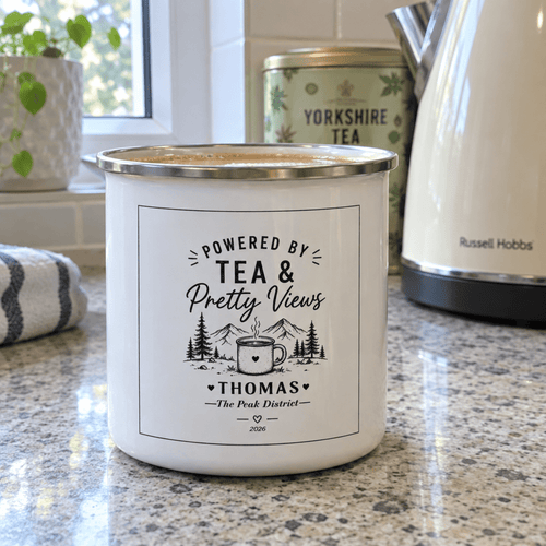 White enamel camping mug with silver rim, “Powered by Tea & Pretty Views” printed label.