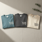 Crewneck Birthday Energy tees in teal, graphite, and ivory with front graphic
