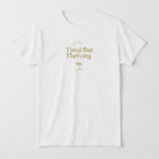 White T-shirt with gold “Tired But Thriving” graphic and leaf accents