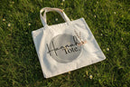 White cotton tote bag with circular “Hannah’s Tote” print and small botanical sprig