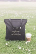 Black tote bag with “caffeine chaos catastrophe” design on grass with coffee cup nearby.