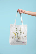 White canvas tote bag with light handle straps and botanical flower print, cursive "Anna"
