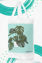 White tote bag with black handles and green monstera leaf print on teal panel