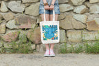 White canvas tote bag with tropical print and teal circular lettering on front.