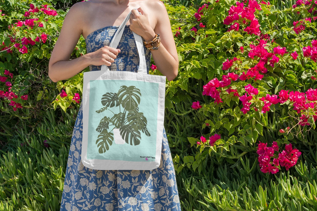 White tote bag with black handles and green monstera leaf print in garden scene