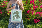 White tote bag with black handles and green monstera leaf print in garden scene