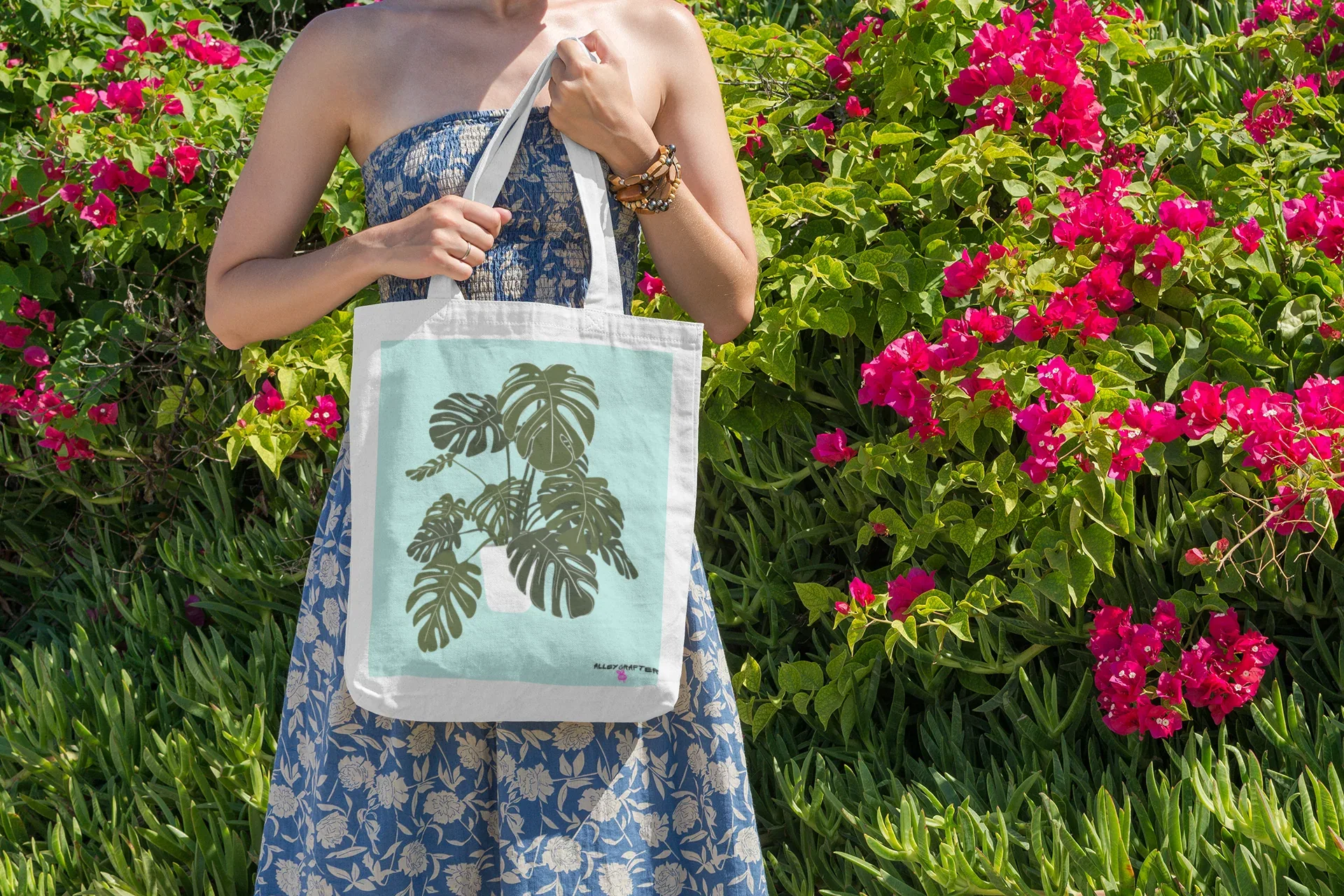 White tote bag with black handles and green monstera leaf print in garden scene