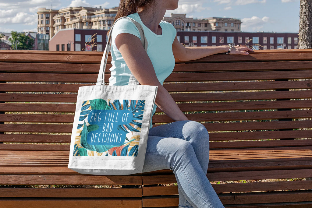White canvas tote bag with tropical leaf graphic and teal “bad decisions” text.