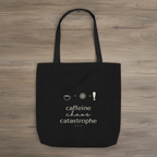 Black tote bag on wood surface with “caffeine chaos catastrophe” text and icons.