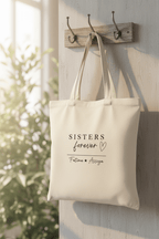 Cream canvas tote bag hanging on wall hook, front shows “SISTERS forever”