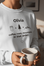 Close-up of white personalized sweatshirt with name Olivia and coffee, trees, mountains