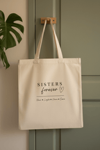 Cream tote bag hanging from wooden hooks, front shows “SISTERS forever”