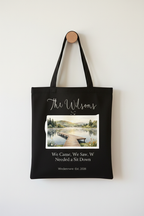 Black canvas tote bag on a white wall, with a printed lakeside boardwalk scene.